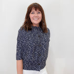 A smiling woman with brown hair and bangs stands against a plain white wall, wearing a navy blue and white textured short-sleeve button-front top with white pants.