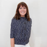 A smiling woman with brown hair and bangs stands against a plain white wall, wearing a navy blue and white textured short-sleeve button-front top with white pants.