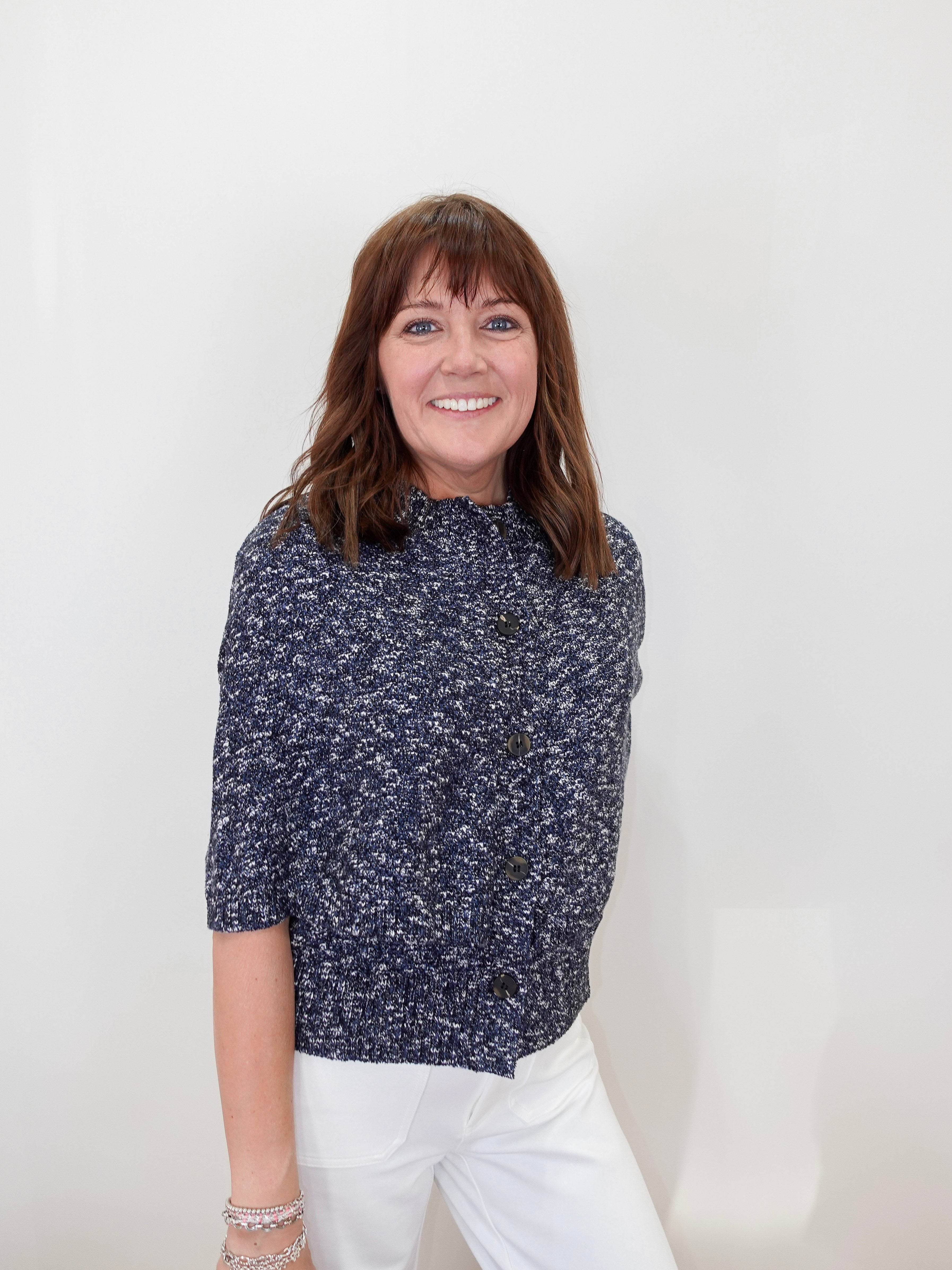 A smiling woman with brown hair and bangs stands against a plain white wall, wearing a navy blue and white textured short-sleeve button-front top with white pants.