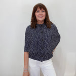 A woman posing in a navy blue and white micro-patterned short-sleeve blouse and white trousers, holding a black handbag against a plain white background.