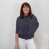 A woman posing in a navy blue and white micro-patterned short-sleeve blouse and white trousers, holding a black handbag against a plain white background.