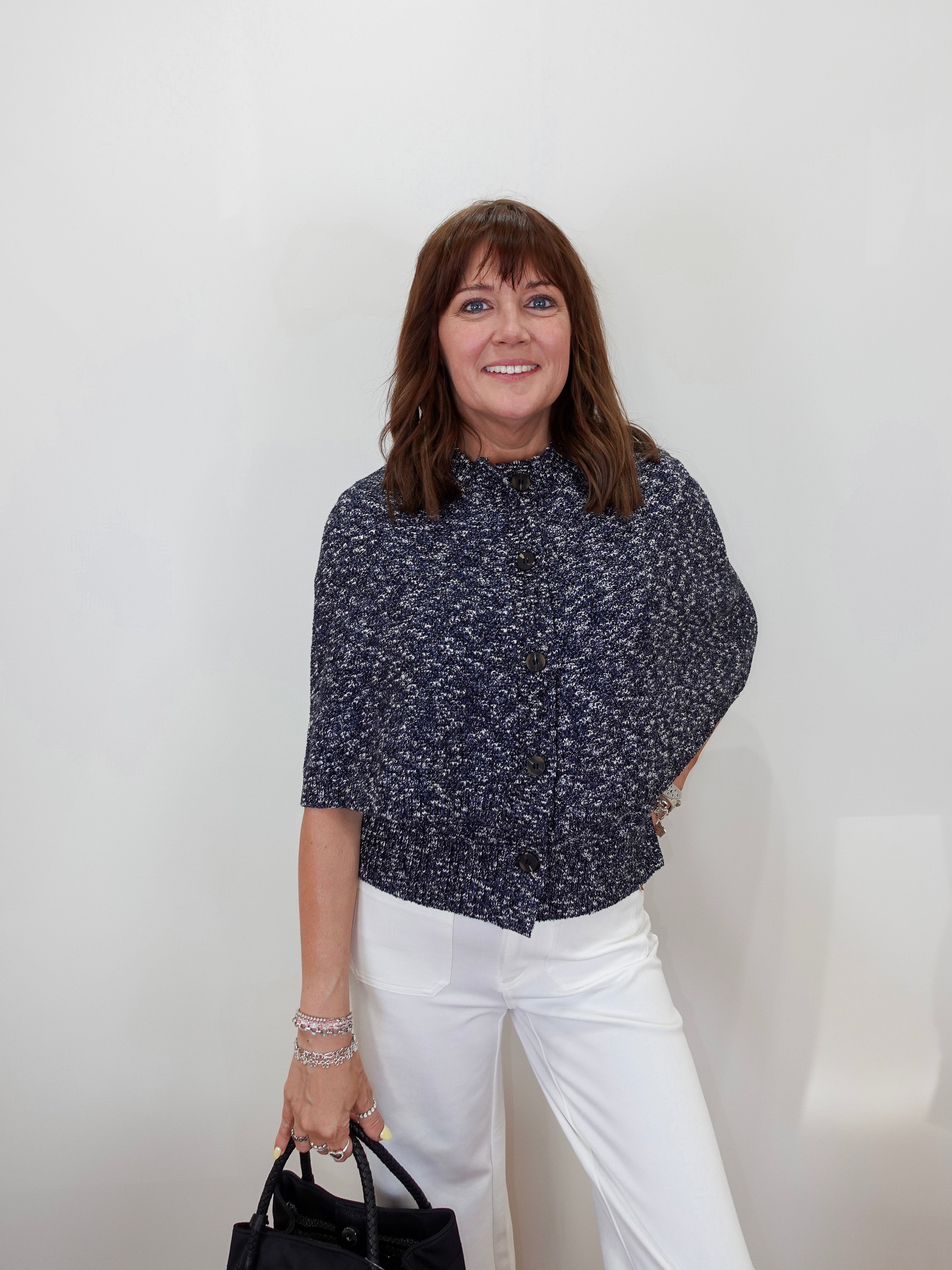 A woman posing in a navy blue and white micro-patterned short-sleeve blouse and white trousers, holding a black handbag against a plain white background.