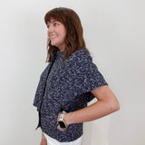 A woman wearing a navy blue short-sleeve top with a white speckled pattern and white pants, standing against a plain white wall.