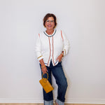 Woman wearing a white sweater with red accents and blue jeans, holding a yellow handbag against a white wall.