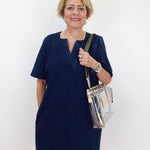 An older woman with short blonde hair stands against a plain white background wearing a short-sleeved navy blue dress and holding a clear plastic shoulder bag with gold trim.