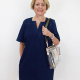 An older woman with short blonde hair stands against a plain white background wearing a short-sleeved navy blue dress and holding a clear plastic shoulder bag with gold trim.