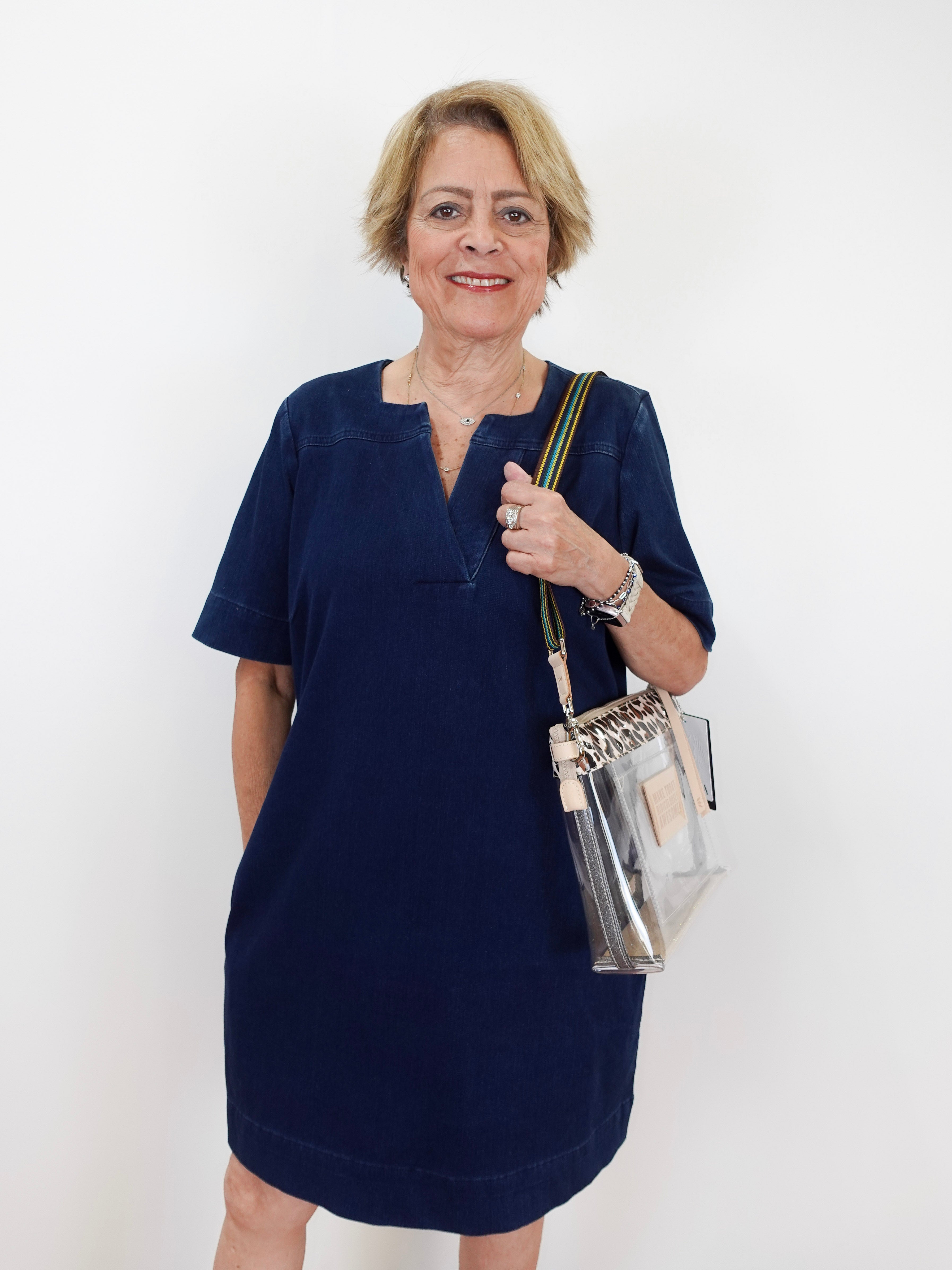 An older woman with short blonde hair stands against a plain white background wearing a short-sleeved navy blue dress and holding a clear plastic shoulder bag with gold trim.
