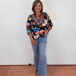 A woman wearing a black floral long-sleeve blouse with colorful trim, light blue flared jeans, and dark brown open-toe heels, standing against a plain white wall.