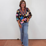 A woman wearing a black floral long-sleeve blouse with colorful trim, light blue flared jeans, and dark brown open-toe heels, standing against a plain white wall.