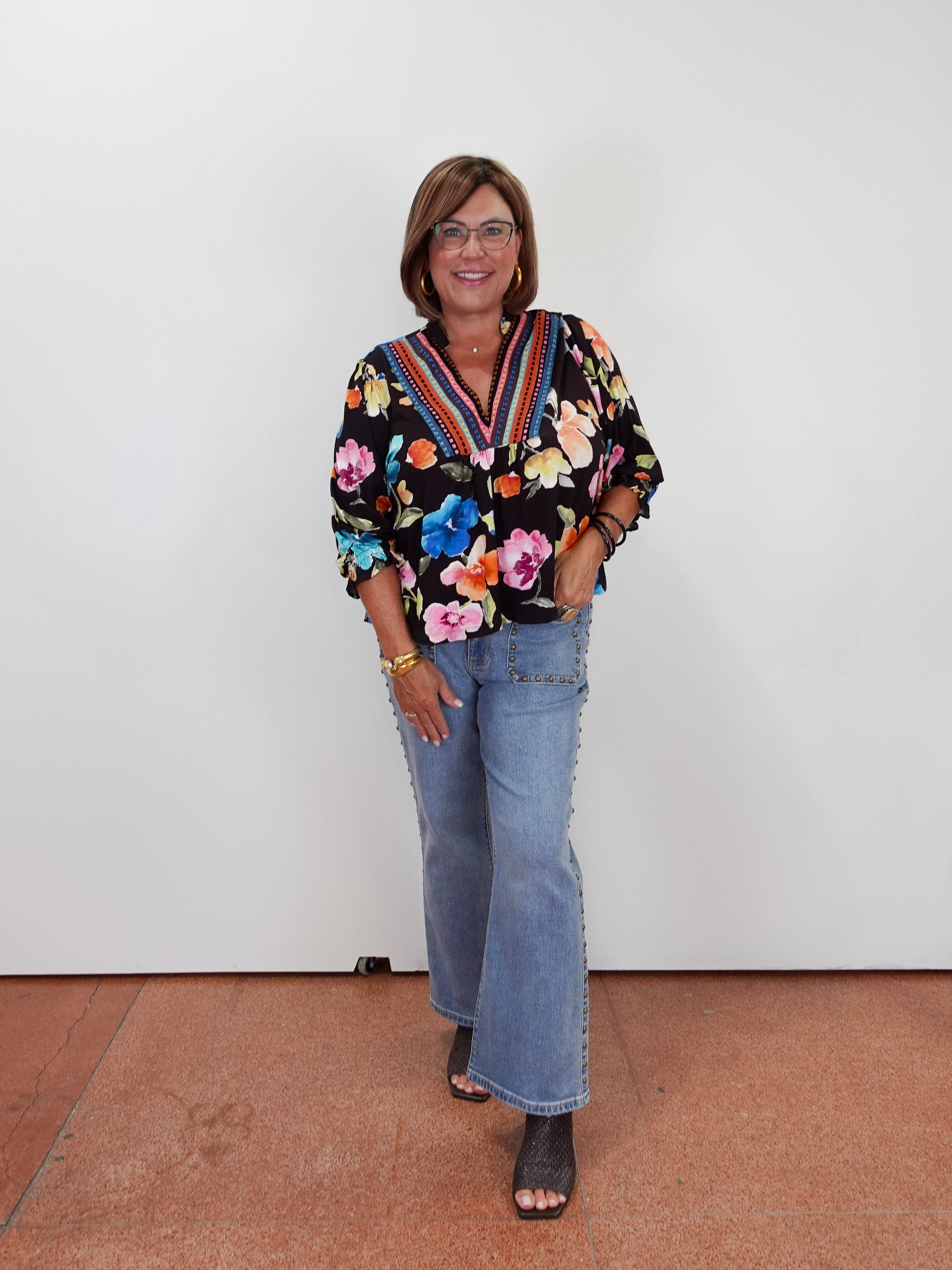 A woman wearing a black floral long-sleeve blouse with colorful trim, light blue flared jeans, and dark brown open-toe heels, standing against a plain white wall.