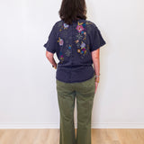 Person wearing a dark blue shirt with floral embroidery and green pants against a white wall.