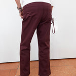 A person shown from the back wearing burgundy straight-leg pants, a white top, and black sandals, standing against a white wall with terracotta tiled flooring.