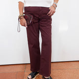 A person wearing straight-leg burgundy pants, a white long-sleeve top, and black platform sandals, holding a small burgundy wristlet bag against a white wall.