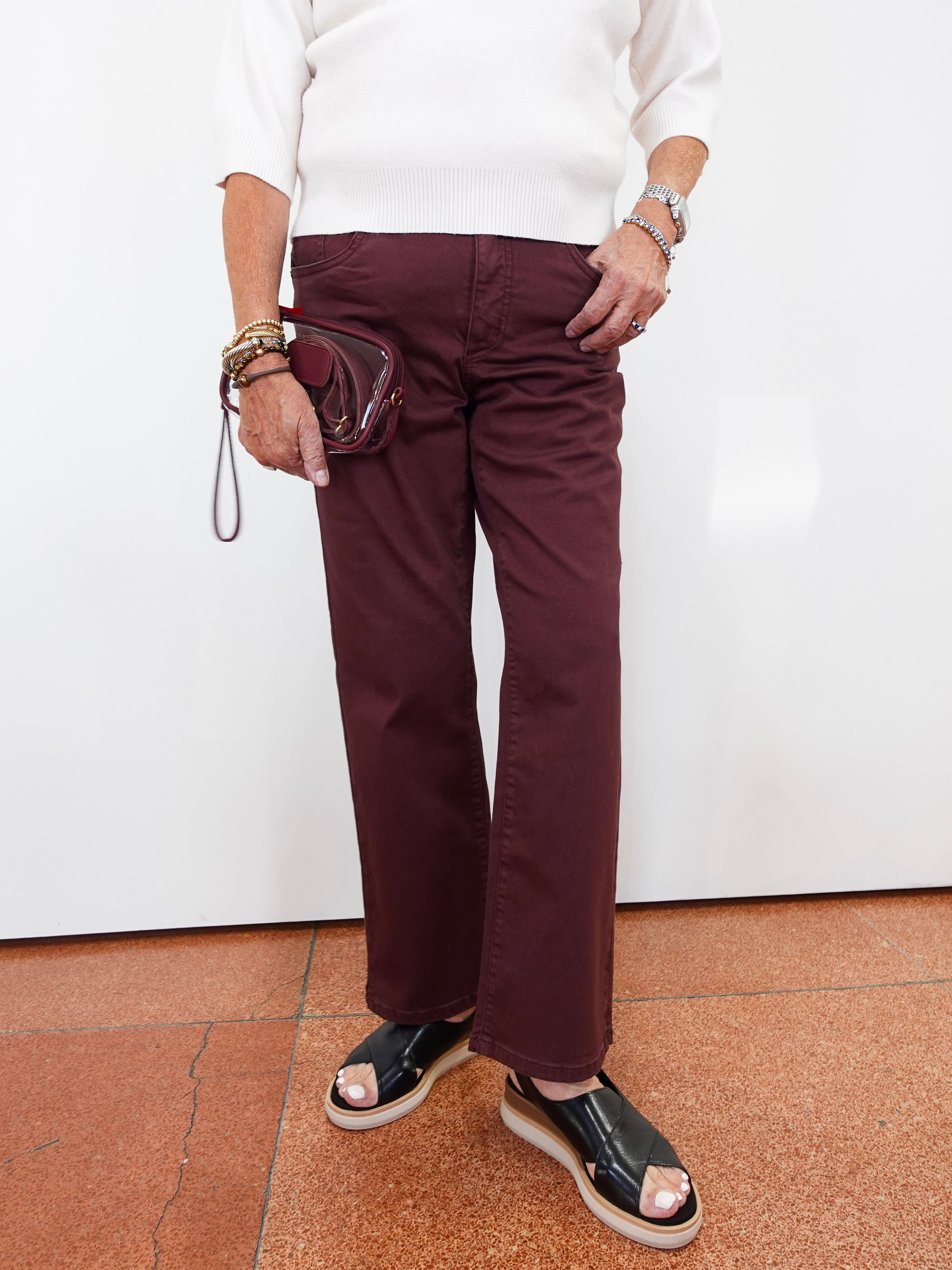 A person wearing straight-leg burgundy pants, a white long-sleeve top, and black platform sandals, holding a small burgundy wristlet bag against a white wall.