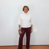 A woman stands against a plain white wall wearing a white mock-neck knit top, burgundy wide-leg pants, and black sandals, holding a small dark clutch.