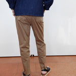 A person shown from the back wearing tan chino pants, a dark blue denim jacket, and black sandals with white soles, standing against a white wall on a terracotta-colored tiled floor.