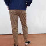 A person shown from the back wearing tan chino pants, a dark blue denim jacket, and black sandals with white soles, standing against a white wall on a terracotta-colored tiled floor.