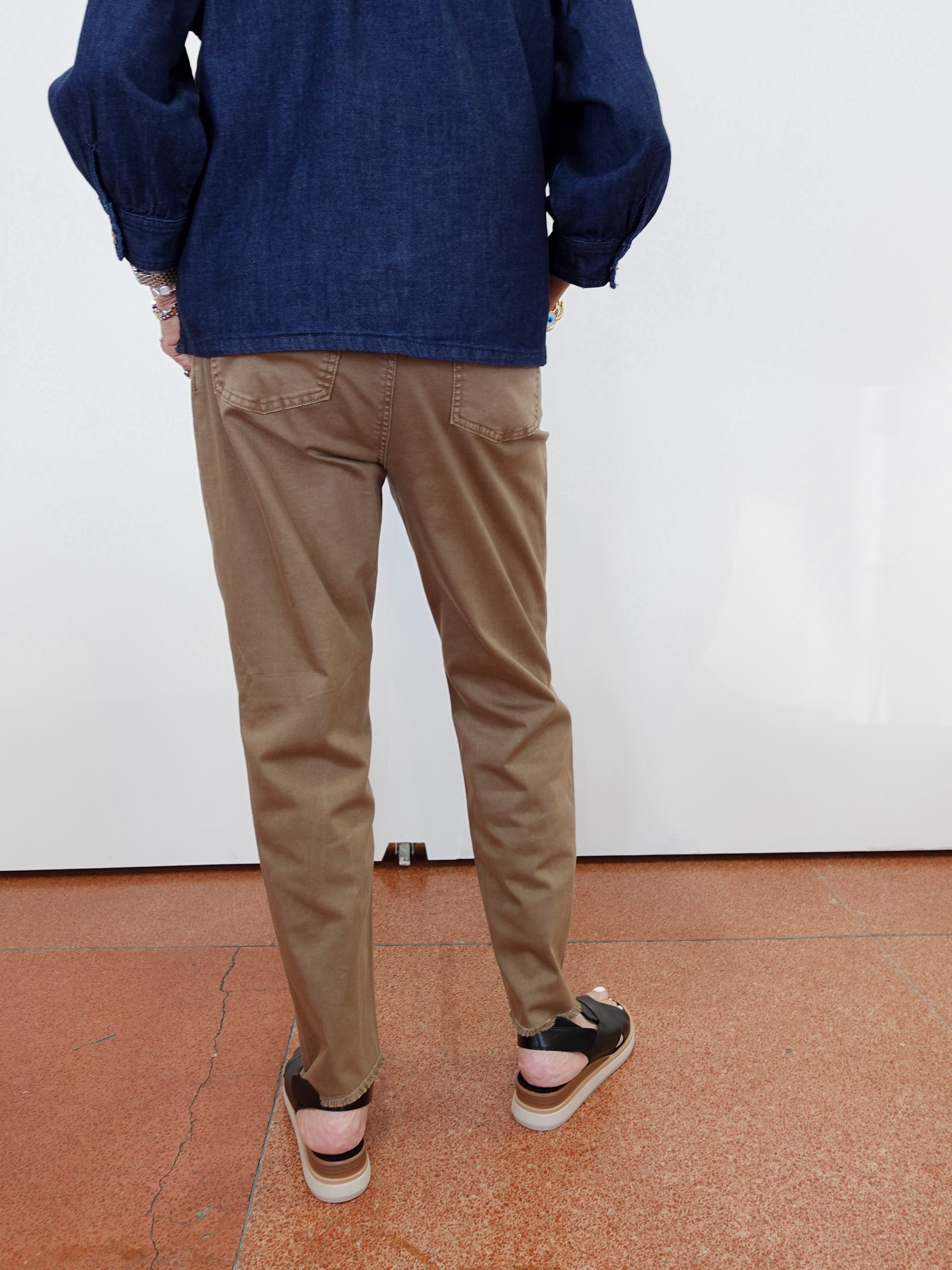 A person shown from the back wearing tan chino pants, a dark blue denim jacket, and black sandals with white soles, standing against a white wall on a terracotta-colored tiled floor.