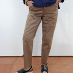 A person wearing slim-fit tan chino pants, a dark blue long-sleeve shirt, and black slide sandals, standing against a white wall with terracotta tiled flooring.