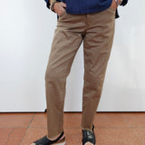 A person wearing slim-fit tan chino pants, a dark blue long-sleeve shirt, and black slide sandals, standing against a white wall with terracotta tiled flooring.