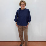 A woman stands against a plain white wall wearing a dark navy blue long-sleeve top, tan pants, and black sandals.