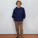A woman stands against a plain white wall wearing a dark navy blue long-sleeve top, tan pants, and black sandals.
