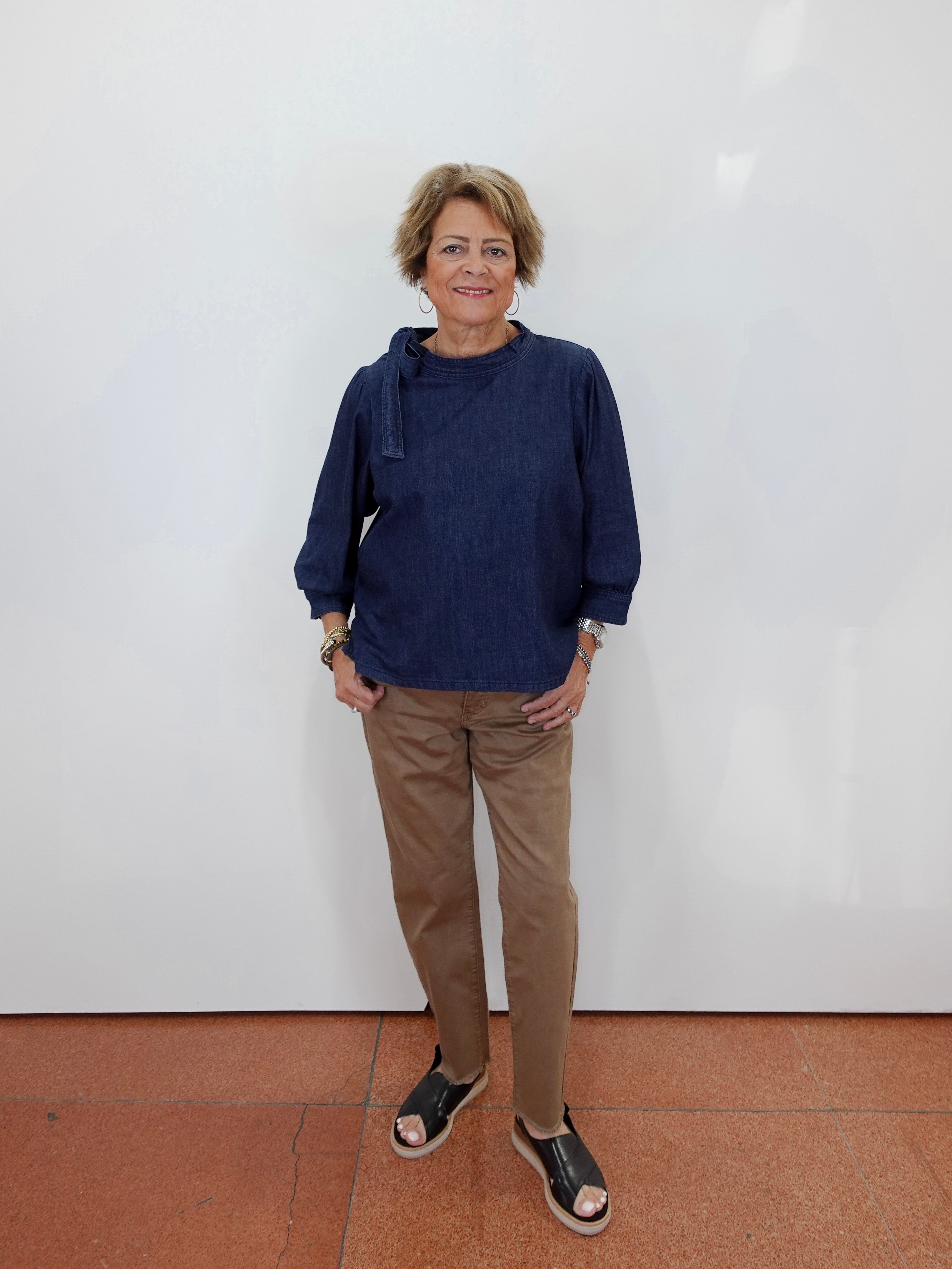 A woman stands against a plain white wall wearing a dark navy blue long-sleeve top, tan pants, and black sandals.