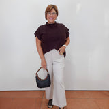 A woman stands against a plain white wall wearing a dark plum mock-neck top, off-white wide-leg pants, and black sandals, while holding a black handbag.