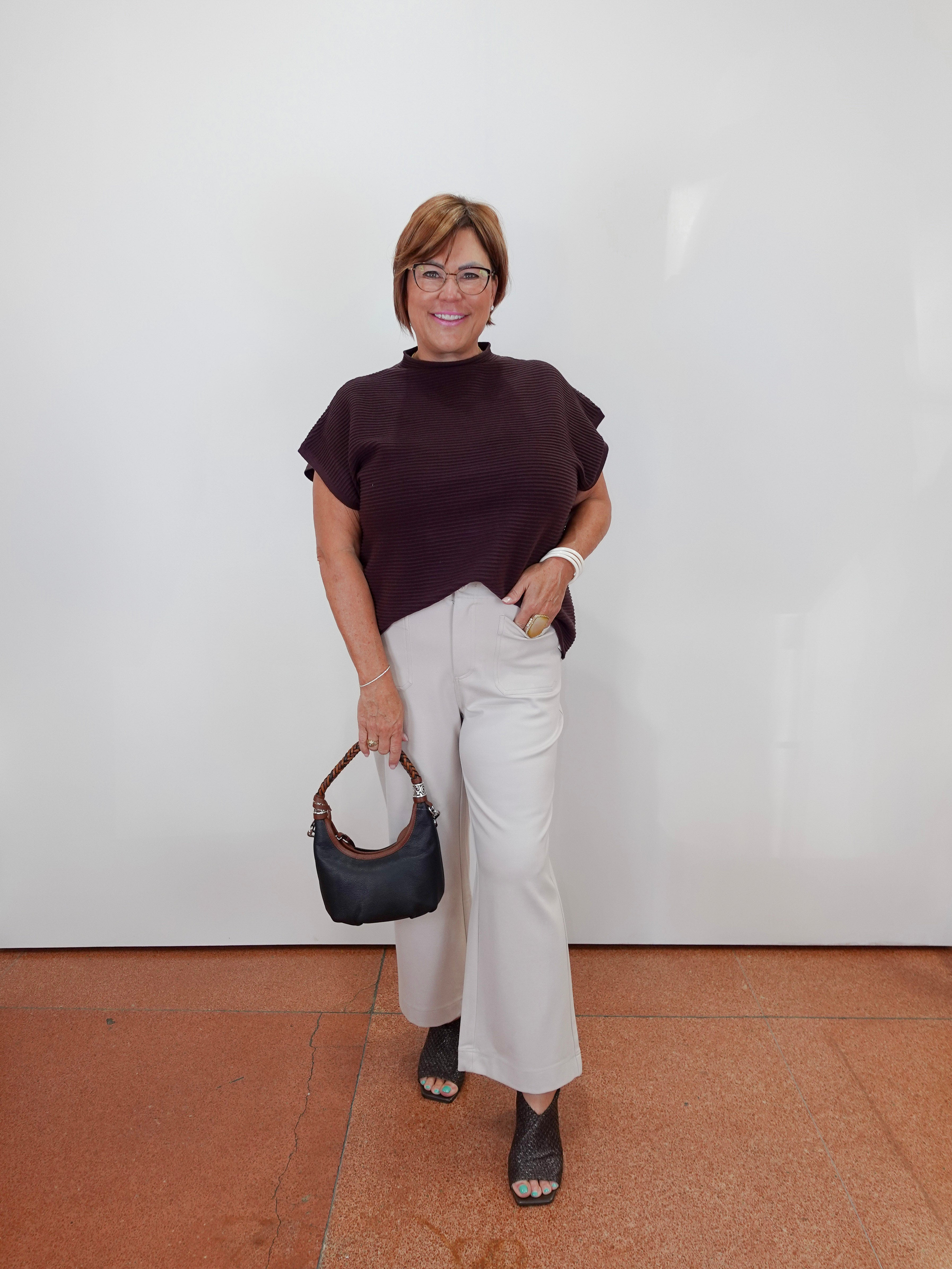 A woman stands against a plain white wall wearing a dark plum mock-neck top, off-white wide-leg pants, and black sandals, while holding a black handbag.