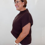 A woman with short brown hair and glasses, wearing a dark brown ribbed short-sleeve mock neck top and white pants, standing in profile against a plain white wall.