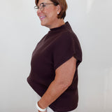 A woman with short brown hair and glasses, wearing a dark brown ribbed short-sleeve mock neck top and white pants, standing in profile against a plain white wall.