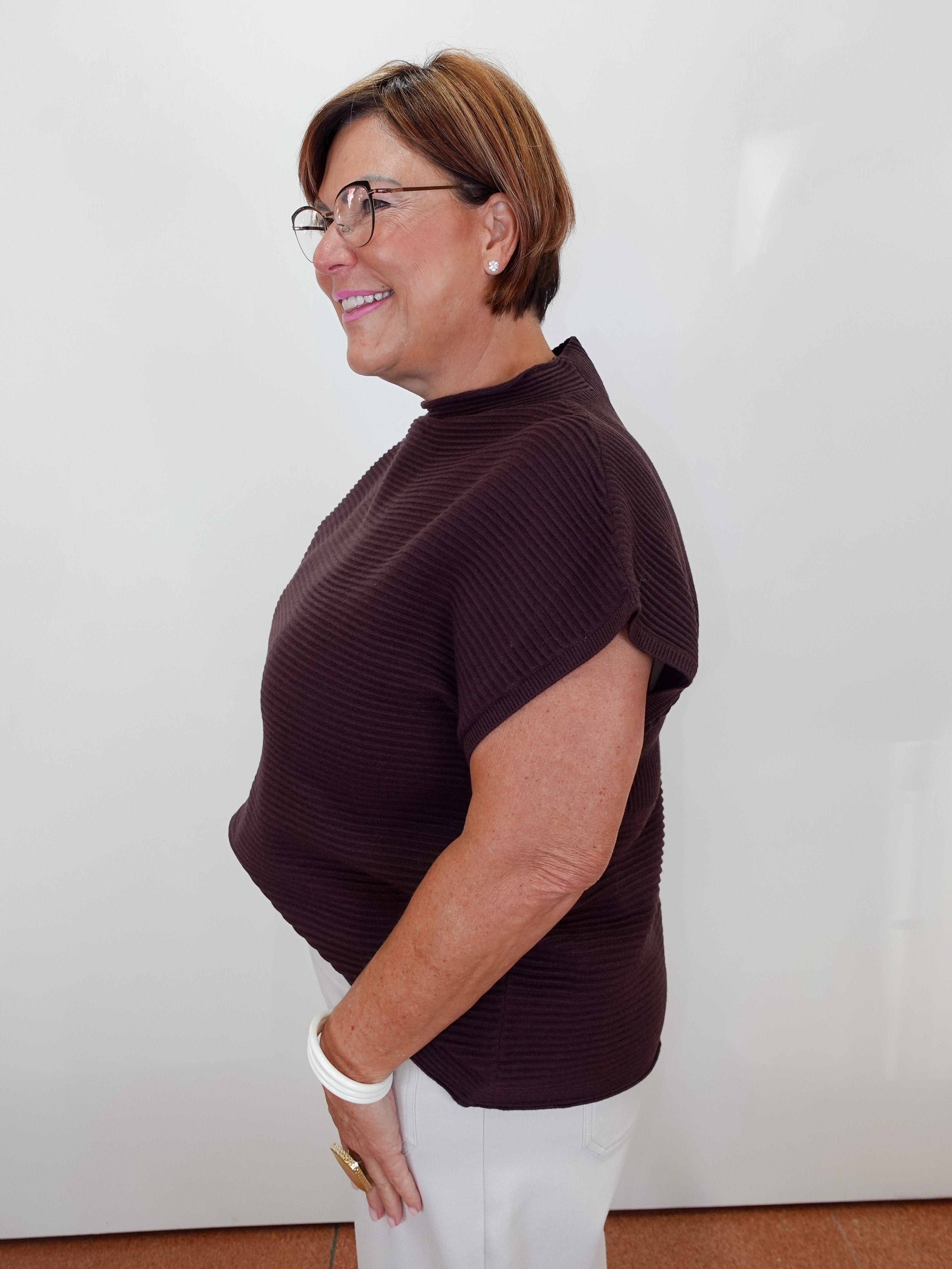 A woman with short brown hair and glasses, wearing a dark brown ribbed short-sleeve mock neck top and white pants, standing in profile against a plain white wall.