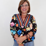 A smiling woman with glasses and short brown hair wearing a black long-sleeve blouse with a vibrant multicolor floral print and embroidered V-neck trim, paired with light blue jeans, standing against a plain white background.