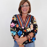 A smiling woman with glasses and short brown hair wearing a black long-sleeve blouse with a vibrant multicolor floral print and embroidered V-neck trim, paired with light blue jeans, standing against a plain white background.