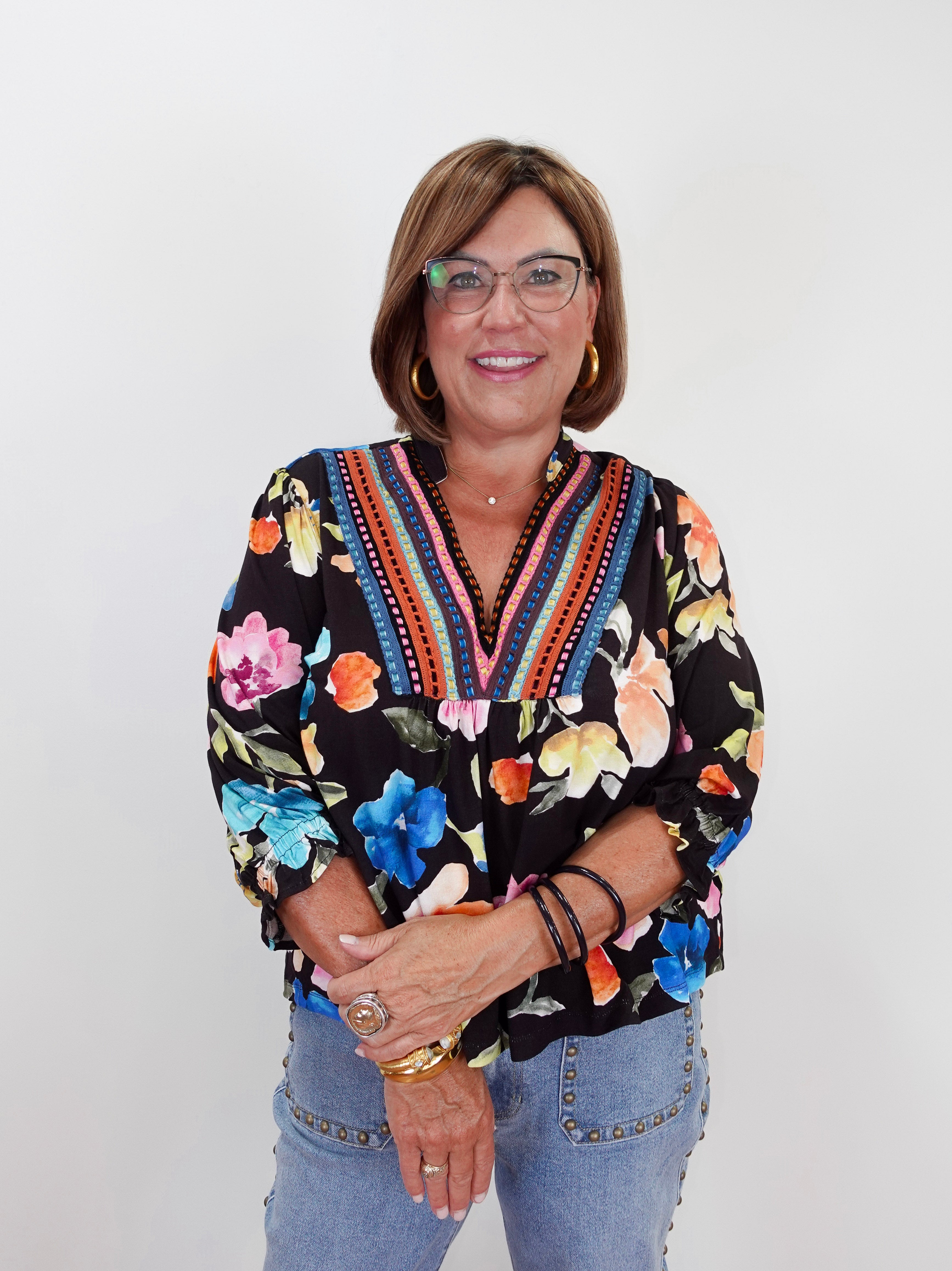 A smiling woman with glasses and short brown hair wearing a black long-sleeve blouse with a vibrant multicolor floral print and embroidered V-neck trim, paired with light blue jeans, standing against a plain white background.
