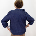 A person viewed from behind wearing a dark indigo blue denim shirt with rolled-up sleeves and tan pants, standing against a plain white wall.