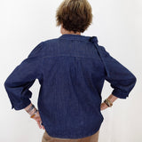 A person viewed from behind wearing a dark indigo blue denim shirt with rolled-up sleeves and tan pants, standing against a plain white wall.