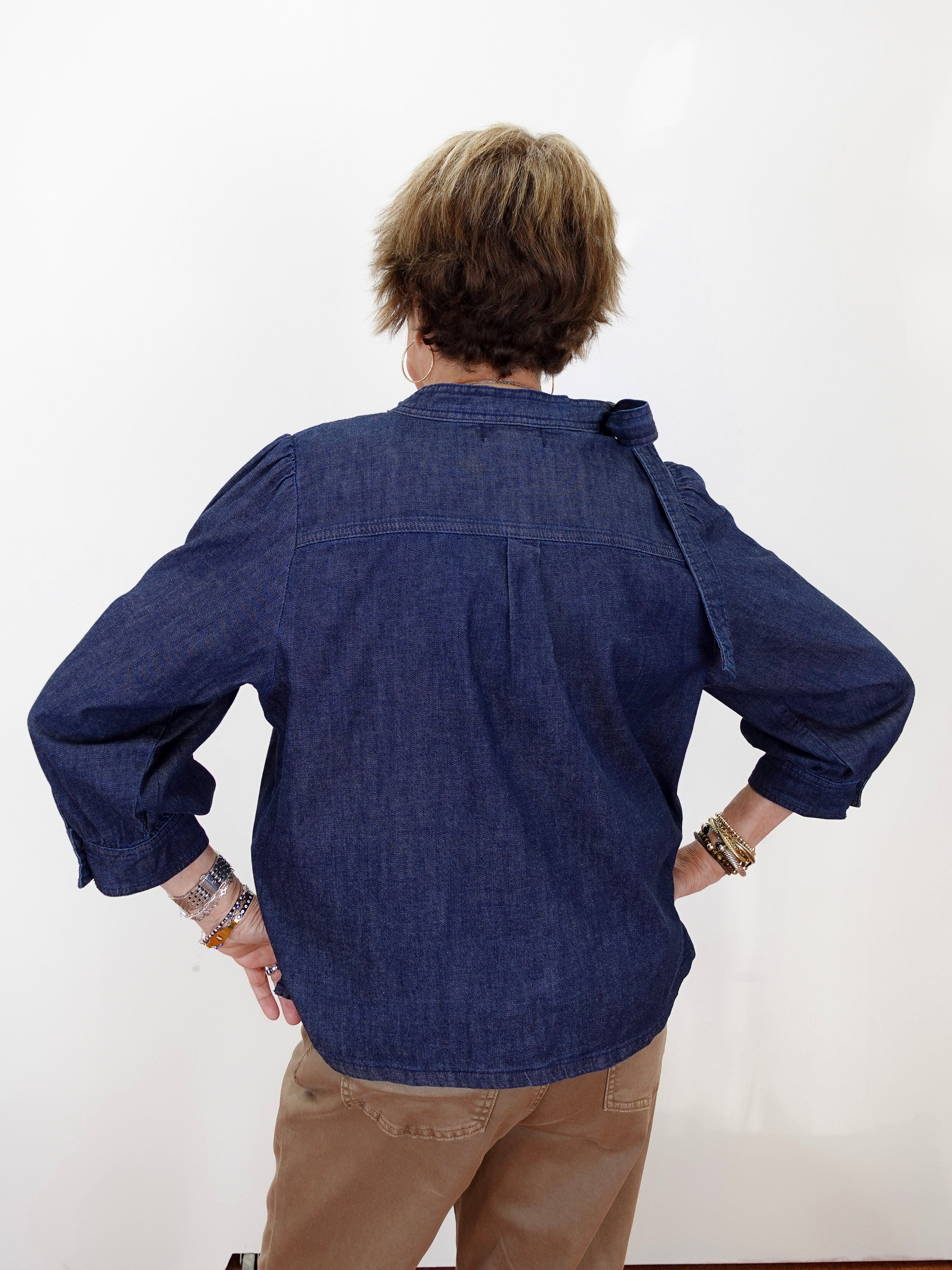 A person viewed from behind wearing a dark indigo blue denim shirt with rolled-up sleeves and tan pants, standing against a plain white wall.