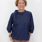 A woman with short blonde hair wearing a dark indigo blue long-sleeve top and tan pants, standing against a plain white background.