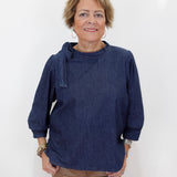 A woman with short blonde hair wearing a dark indigo blue long-sleeve top and tan pants, standing against a plain white background.