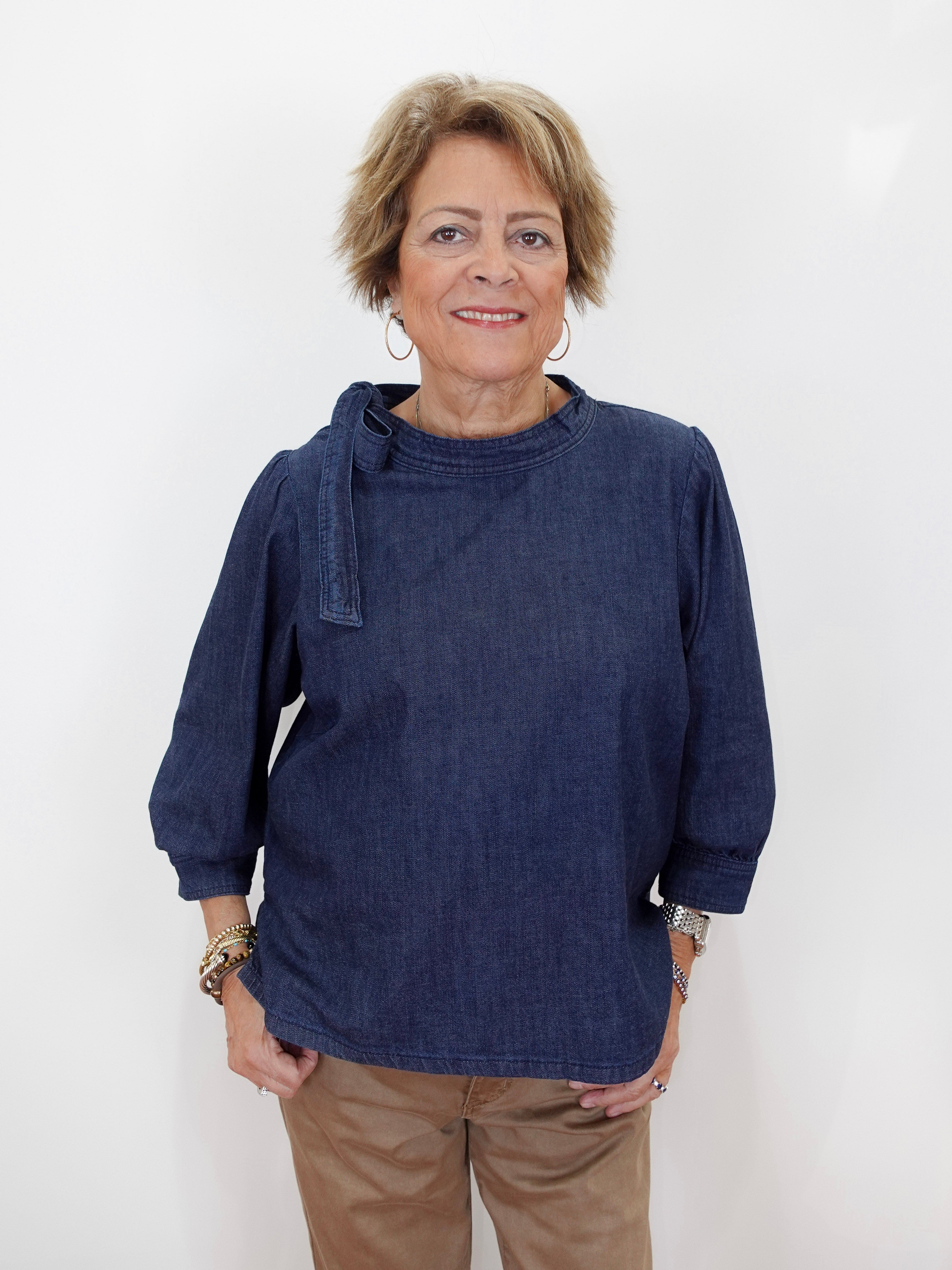 A woman with short blonde hair wearing a dark indigo blue long-sleeve top and tan pants, standing against a plain white background.