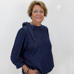 A woman with short blonde hair wearing a dark indigo denim long-sleeve top and tan pants, standing against a plain white background.