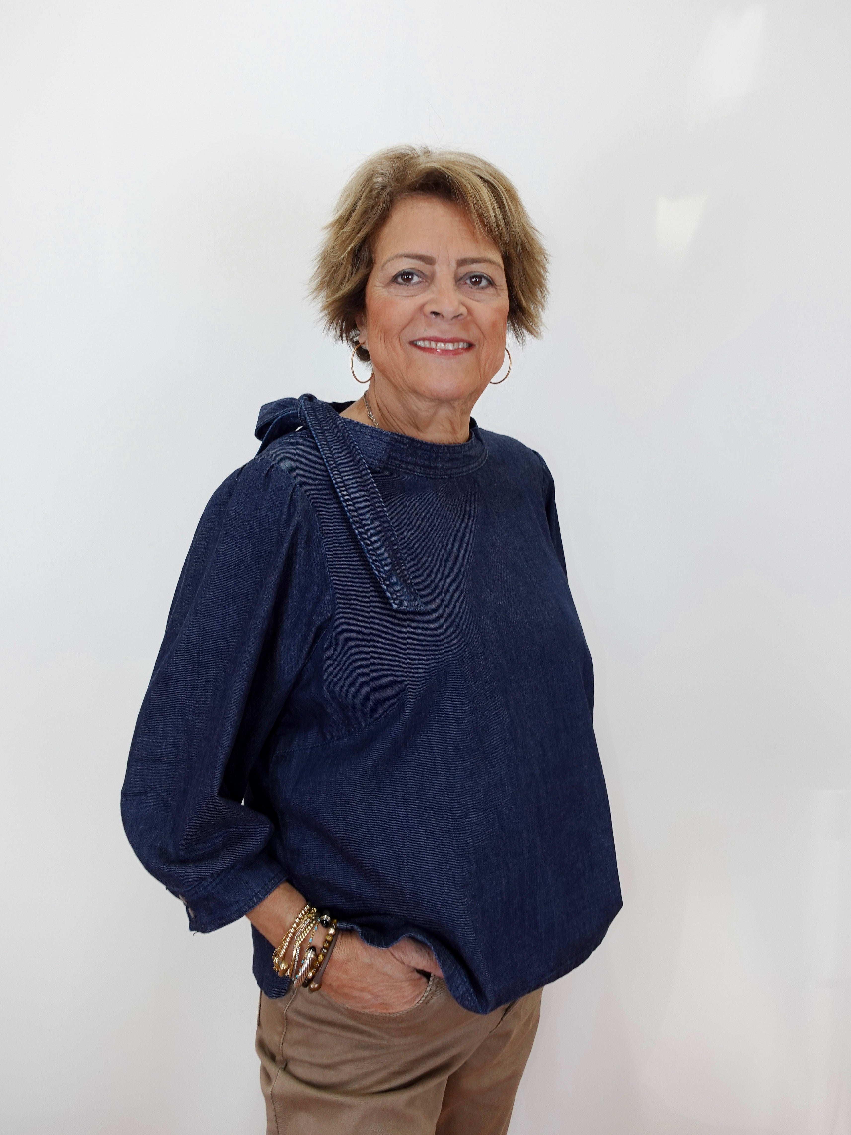 A woman with short blonde hair wearing a dark indigo denim long-sleeve top and tan pants, standing against a plain white background.