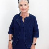 An older woman with short white hair wearing a navy blue corduroy jacket and matching skirt, accessorized with a black and white patterned headband, standing against a plain white background.