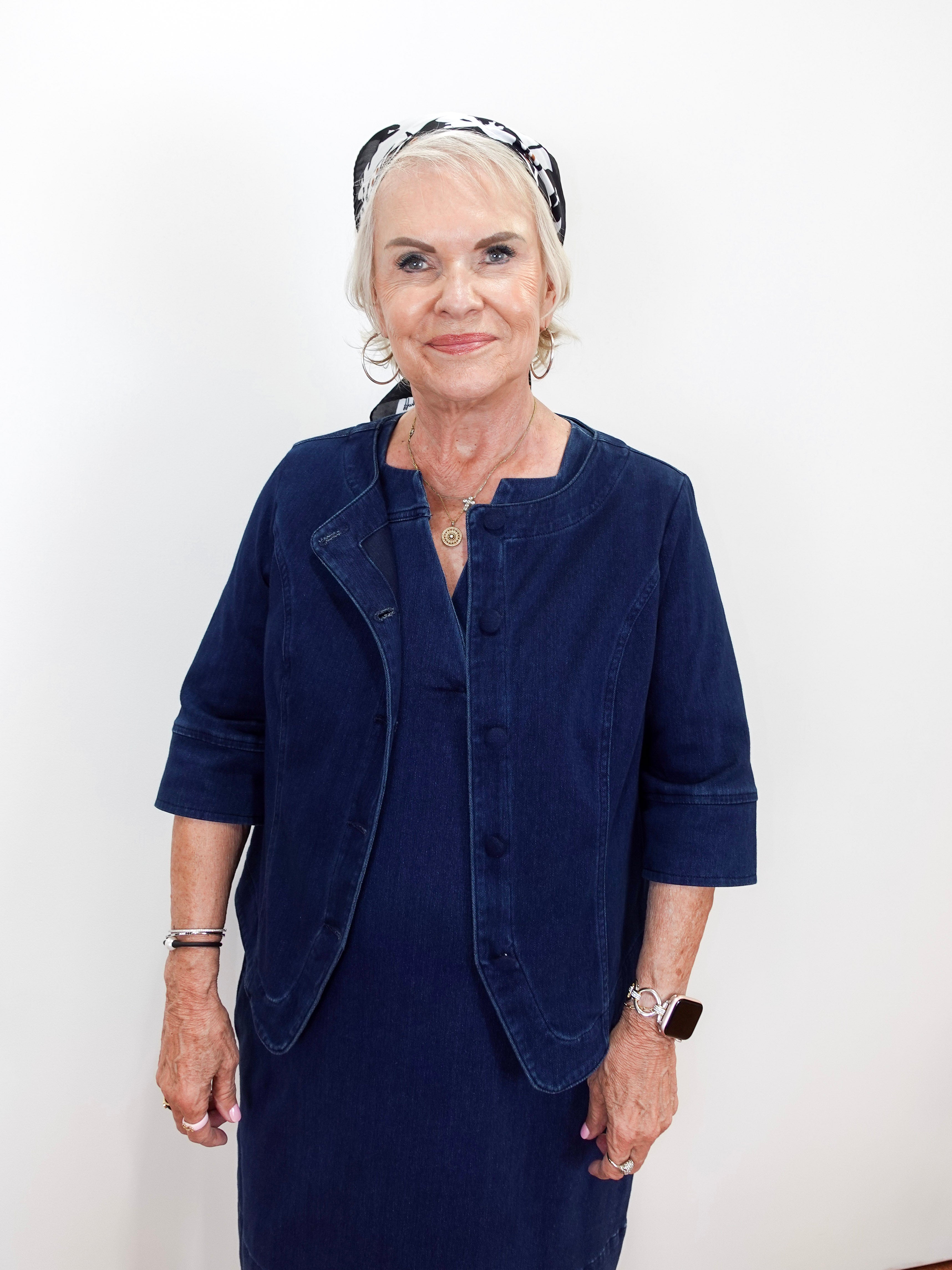 An older woman with short white hair wearing a navy blue corduroy jacket and matching skirt, accessorized with a black and white patterned headband, standing against a plain white background.