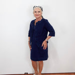 An older woman stands against a plain white wall wearing a dark navy blue dress with a matching jacket-style overlay, beige sandals, and a black-and-white patterned headband.