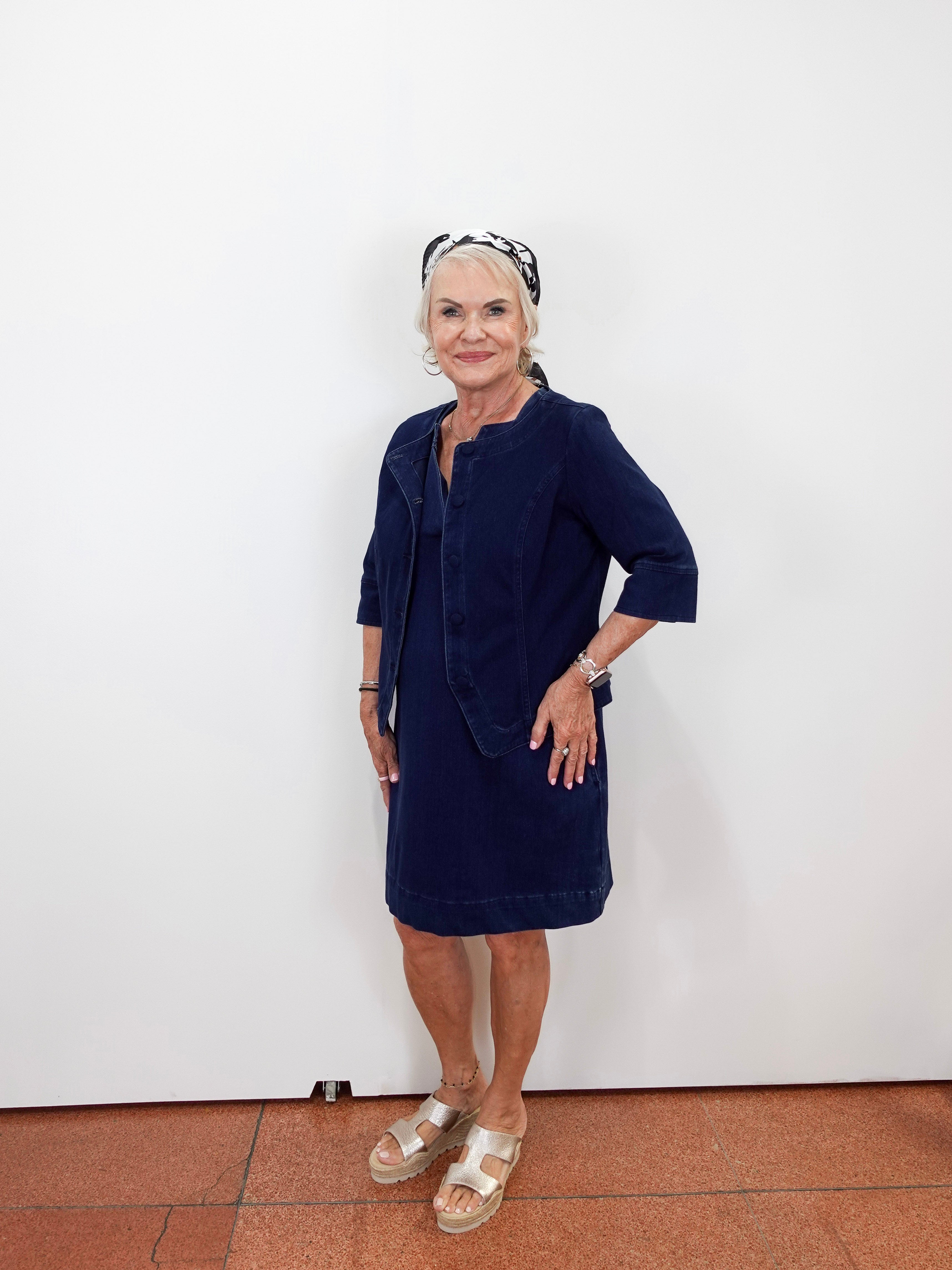 An older woman stands against a plain white wall wearing a dark navy blue dress with a matching jacket-style overlay, beige sandals, and a black-and-white patterned headband.