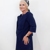 An older woman with blonde hair tied back in a patterned headscarf, wearing a dark navy blue corduroy dress with a matching cropped jacket, posing against a plain white wall.