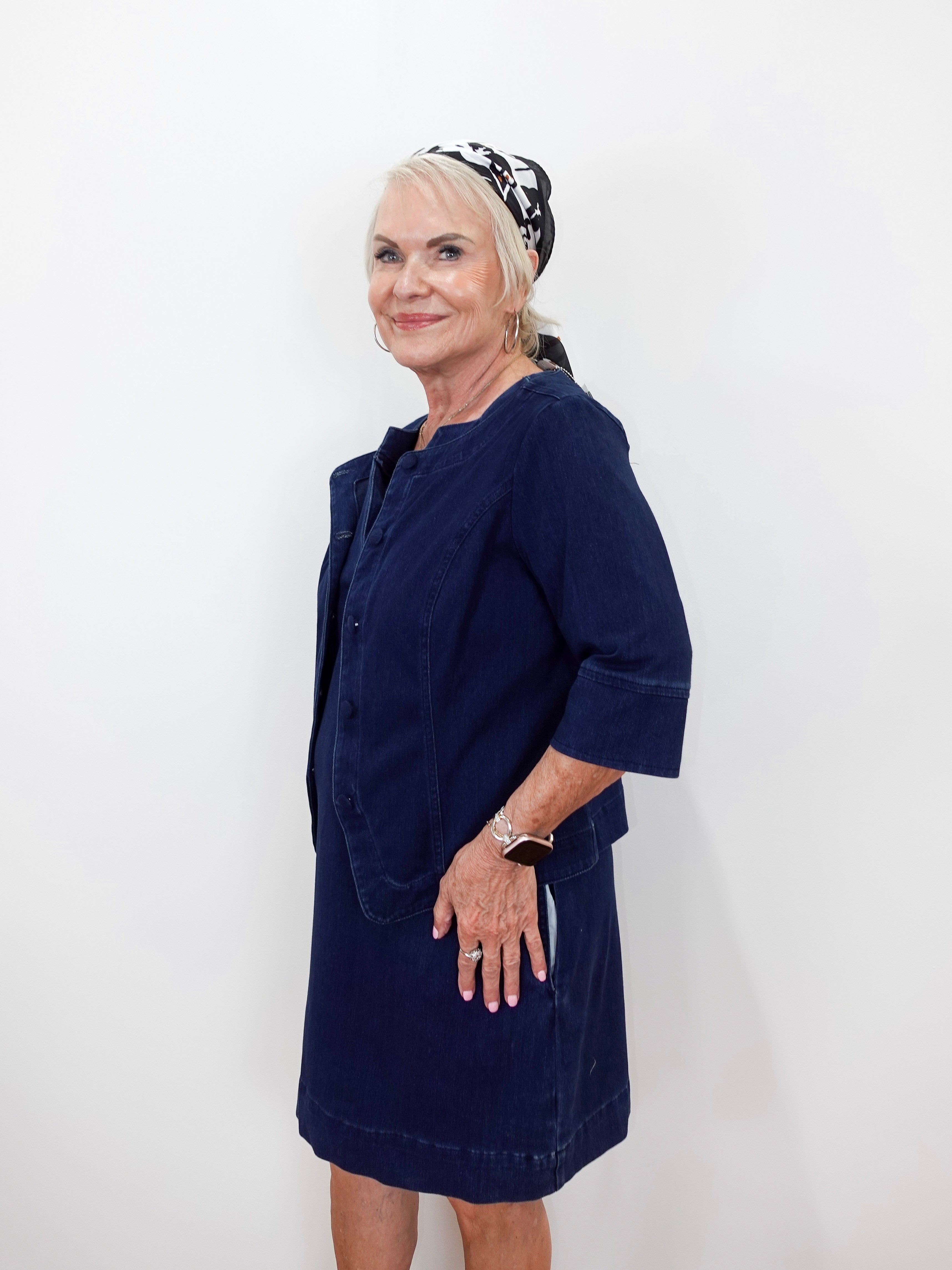 An older woman with blonde hair tied back in a patterned headscarf, wearing a dark navy blue corduroy dress with a matching cropped jacket, posing against a plain white wall.