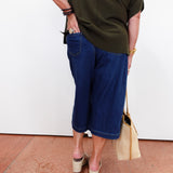 A person shown from the back wearing dark blue cropped denim shorts, an olive green top, and beige wedge sandals, holding a beige tote bag against a white wall.